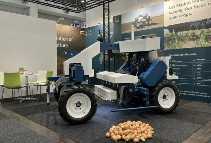 A blue and white Ekobot agricultural robot is displayed in an exhibition booth, with a pile of onions on the floor in front of it. Posters in the background read 